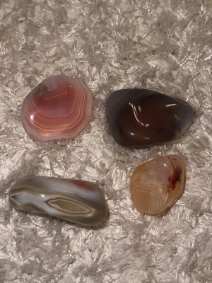 Fyra polerade agatstenar - Säljer fyra polerade agatstenar i olika färger och mönster. Stenarna har mjuka, ovala former och varierar i nyanser av rosa, brun, beige och grått med naturliga band och ådringar. Perfekta som dekoration eller till smyckestillverkning.