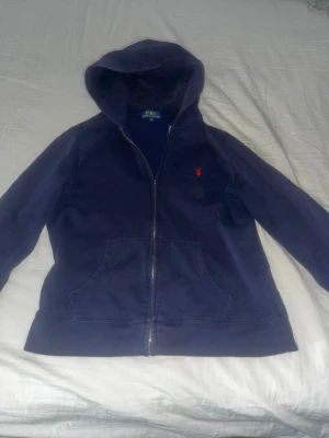 Marinblå hoodie från Polo Ralph Lauren - Snygg marinblå hoodie från Polo Ralph Lauren med dragkedja framtill och klassisk röd logga broderad på bröstet. Hoodien har huva och två fickor. Tillverkad i mjukt bomullsmaterial för skön känsla. Nackdelen är att en del på dragkedjan har lossnat.  (kolla bild 3) Passar också M skulle jag tro. 