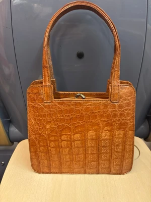 Vintage handväska i äkta krokodilskinn  - Säljer en snygg brun handväska i äkta krokodilskinn. Väskan har ett klassiskt metallspänne upptill och rymmer det viktigaste. Perfekt för dig som gillar vintagevibbar och stilren design. Bredden :20 cm Längden: 15cm  Djup : 7cm