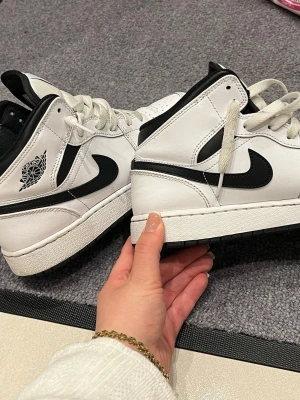 Air Jordan 1 Mid vit/svart - Säljer ett par Air Jordan 1 Mid sneakers i vitt och svart läder. Klassisk svart swoosh på sidan, vita skosnören och svart yttersula. Wings-logga på ankeln och vadderad krage för extra komfort. Perfekta för dig som gillar streetwear och ikonisk design.