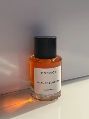 ESSNCE Orange Blossom parfym - En stilren parfymflaska från ESSNCE med doften Orange Blossom. Flaskan är cylindrisk i genomskinligt glas med orangefärgad vätska och svart lock. Etiketten är vit med svart text för en minimalistisk look. Perfekt för dig som gillar fräscha och blommiga dofter.