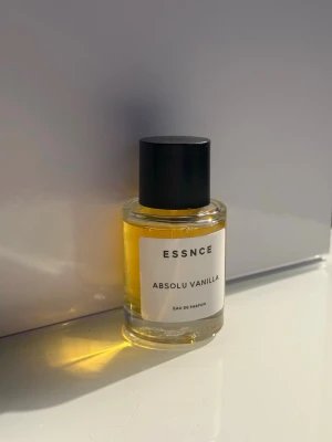 ESSNCE Absolu Vanilla Eau de Parfum - En stilren parfymflaska från ESSNCE med doften Absolu Vanilla. Flaskan är cylindrisk i genomskinligt glas med svart lock och etikett i vitt med svart text. Innehållet är gyllengult och flaskan har en modern, minimalistisk design.
