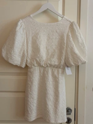 Studentklänning - Supersöt vit kort klänning med puffiga korta ärmar och  rosett i ryggen. Klänningen har en strukturerad, lite krinklad yta och markerad midja. 