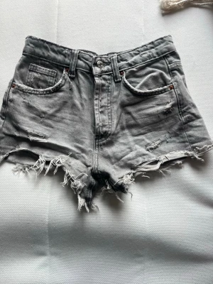 Gråa jeansshorts med slitningar - Säljer ett par gråa jeansshorts med hög midja och råa, fransiga kanter. Shortsen har slitna detaljer framtill och bakfickor. Klassisk femficksmodell i denim med knappgylf. Perfekta för en avslappnad och trendig look.