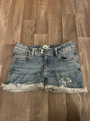 Blå jeansshorts - Säljer ett par blå jeansshorts från 157 med fransig, rå kant och slitningar framtill. De är klippta av mig själv. Klassisk femficksmodell med knappgylf och bälteshällor. Perfekta för sommaren och ger en avslappnad vibe.