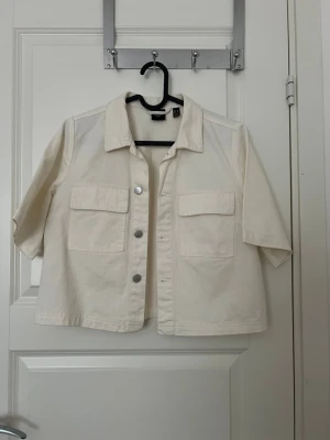 Vit kort jeansjacka från Vero Moda - Stilren vit jeansjacka från Vero Moda med kort, boxig passform och trekvartsärmar. Jackan har två stora bröstfickor med lock och silverfärgade knappar framtill. Tillverkad i kraftigt bomullstyg, perfekt för en trendig och avslappnad look.
