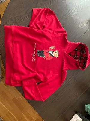 Röd hoodie Polo Ralph Lauren XL - Röd hoodie från Polo Ralph Lauren med ikonisk björn-tryck på bröstet och rutigt foder i huvan. Klassisk passform med långa ärmar och mjukt material. Perfekt för dig som vill sticka ut med en snygg och bekväm look.