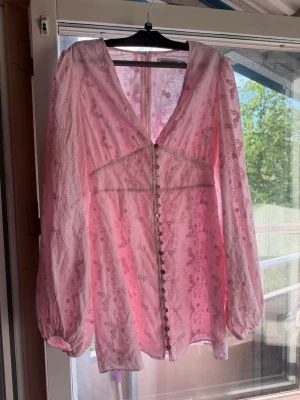 Rosa klänning från Adoore - Supersöt rosa kortklänning från Adoore med blommigt mönster, v-ringning och långa puffärmar. Klänningen har dekorativa knappar framtill. Är liten i storleken så skulle säga att den är mer M i storleken.