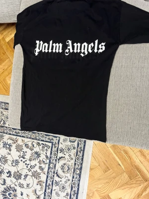 Svart Palm Angels t-shirt - Säljer en svart t-shirt från Palm Angels med stort vitt logotryck på ryggen och mindre logga vid halsen fram. Klassisk passform och rund hals. Perfekt för dig som gillar streetwear och vill ha en stilren look.