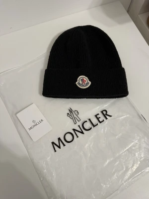 Svart Moncler mössa i ull - Säljer en svart ribbstickad Moncler mössa i ull med uppvikt kant och klassisk Moncler-logga framtill. Mössan är enkel men stilren och passar perfekt till kalla dagar. Kommer med originalpåse och tag.