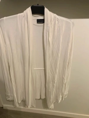 Vit öppen cardigan från KappAhl - Säljer en vit, tunn och öppen cardigan från KappAhl. Koftan har lång ärm, draperad framkant och ett mjukt, följsamt tyg som ger ett snyggt fall. Perfekt att slänga över en topp för en chill look.