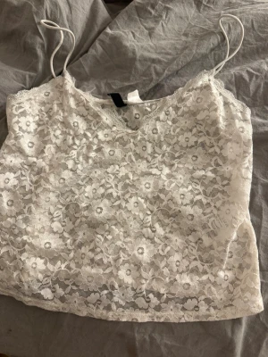 Vit spetslinnetopp med smala band - Säljer en vit spetslinnetopp med smala axelband och blommigt spetsmönster. Toppen är ärmlös och har en croppad passform som passar perfekt till både jeans och kjol. Superfin detalj med spetsen som ger en romantisk vibe.