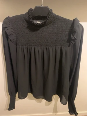 Svart blus från BikBok - Trendig svart blus från BikBok med smockad överdel, volangdetaljer vid axlar och hals samt långa ärmar med smock vid ärmslut. Tillverkad i ett lätt och luftigt tyg som ger en snygg siluett. Perfekt för att lyfta vilken outfit som helst.