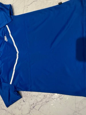 Blå sporttröja från Nike - Snygg blå sporttröja från Nike med vit logga och vit vågig linje över bröstet. Tillverkad i ett lätt och snabbtorkande material som passar perfekt till träning eller match. Klassisk rund hals och korta ärmar för maximal rörelsefrihet.
