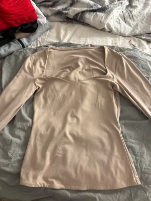 Beige långärmad topp med fyrkantig ringning - Snygg beige långärmad topp med en modern fyrkantig ringning. Toppen har en figurnära passform och är tillverkad i ett mjukt, stretchigt material som känns skönt mot huden. Perfekt att styla med jeans eller kjol för en trendig look.