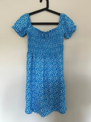 Blå blommig sommarklänning - Söt blå klänning med vita småblommor och korta puffärmar. Klänningen har smock upptill och en lätt utställd kjol. Perfekt för varma dagar och har en ungdomlig, trendig stil. Materialet känns lätt och luftigt.