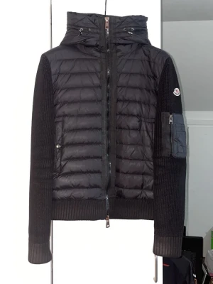 Svart pufferjacka från Moncler - Snygg svart pufferjacka från Moncler med stickade ärmar och vadderad front. Jackan har huva med dragsko, dragkedja framtill och en praktisk ficka med dragkedja på ärmen. Perfekt för kalla vinterdagar och ger en sportig, trendig look. Kvitto och allt ifrån köpet finns