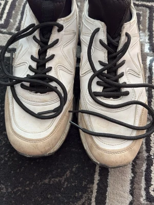 Vita sneakers med svarta detaljer - Säljer ett par vita sneakers med svarta skosnören och svart plös. Skorna har en chunky sula och paneler i syntetmaterial. Passar perfekt till streetwear  och har en modern look.
