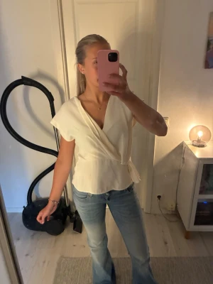 Vit omlottblus med knytband - Snygg vit omlottblus med korta ärmar och knytband i midjan. Blusen har en avslappnad passform och lätt v-ringad halsringning. Perfekt att matcha med jeans för en fräsch look.