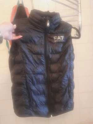 Svart dunväst från EA7 Emporio Armani - Snygg svart dunväst från EA7 Emporio Armani med hög krage och dragkedja framtill. Västen har ett stilrent, quiltat mönster och logga på bröstet. Perfekt för lager på lager och ger en sportig vibe till din outfit.