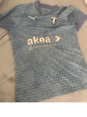 Ljusblå Malmö FF matchtröja Puma - Säljer en ljusblå Malmö FF matchtröja från Puma med geometriskt mönster och klubbmärke på bröstet. Tröjan har korta ärmar, rund hals och sponsorlogga framtill. Perfekt för dig som älskar fotboll och vill stötta laget på match.
