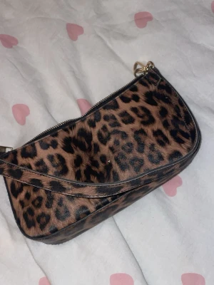 Leopardmönstrad axelväska - Snygg handväska med leopardmönster i brunt och svart. Väskan har en kort axelrem och dragkedja upptill. Materialet ser ut som konstläder med pälsstruktur. Perfekt för dig som vill sticka ut med ett djurmönstrat statement.