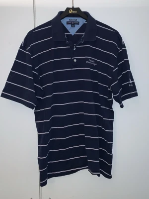Marinblå randig pikétröja Tommy Hilfiger - Snygg marinblå pikétröja från Tommy Hilfiger med vita ränder och broderad text på bröstet. På ärmen finns en fjäril, texten Kapalua och Tommy Hilfigers logga. Klassisk krage och knappar framtill. Perfekt för en avslappnad men stilren look.