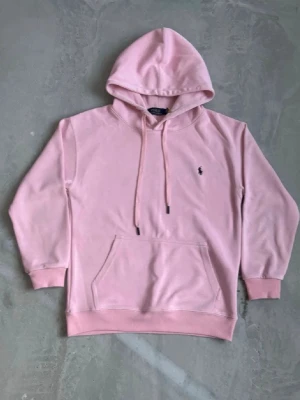 Ljusrosa hoodie från Polo Ralph Lauren - Snygg ljusrosa hoodie från Polo Ralph Lauren med klassisk huva och dragsko. Har en broderad logga på bröstet och en stor magficka framtill. Perfekt för en chill och stilren look. Mjuk bomullskänsla och ribbade muddar vid ärmslut och nederkant.