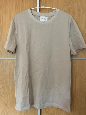 Beige t-shirt från SANDÖ - Säljer en stilren beige t-shirt från SANDÖ i mjuk bomull. T-shirten har klassisk rund halsringning och korta ärmar. Perfekt basplagg att matcha med allt och superbekväm att bära hela dagen.