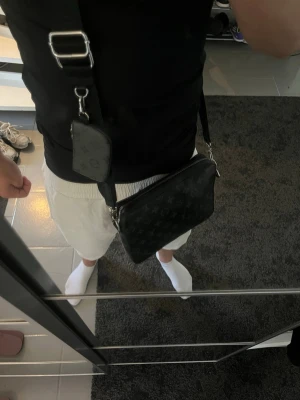 Svart axelväska med monogram - Snygg svart axelväska med justerbar bred rem och diskret monogrammönster. Väskan har en fyrkantig form och en mindre avtagbar ficka på remmen. Perfekt för dig som vill ha en stilren och praktisk accessoar.