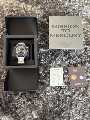 Omega x Swatch MoonSwatch Mercury - Snygg Omega x Swatch MoonSwatch Mission to Mercury med svart urtavla, vita detaljer och grått band. Klockan har tachymeter och flera subdials. Levereras i originalbox med tillhörande manualer och certifikat.