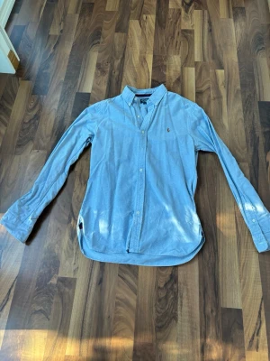 Blå skjorta från Polo Ralph Lauren - Snygg ljusblå skjorta från Polo Ralph Lauren i slim fit-modell. Skjortan har klassisk krage, knappar framtill och lång ärm. Tillverkad i bomull och har den ikoniska lilla loggan broderad på bröstet.