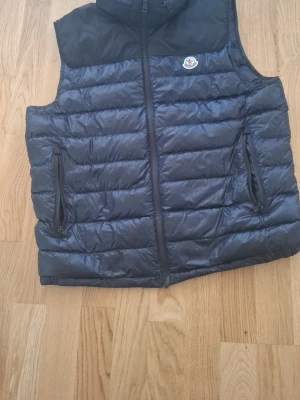 Svart dunväst från Moncler  - Snygg svart dunväst från Moncler med quiltad design och dragkedja framtill. Västen har två praktiska fickor med dragkedja och en diskret Moncler-logga på bröstet. Perfekt för lager-på-lager och håller dig varm under kyliga dagar.