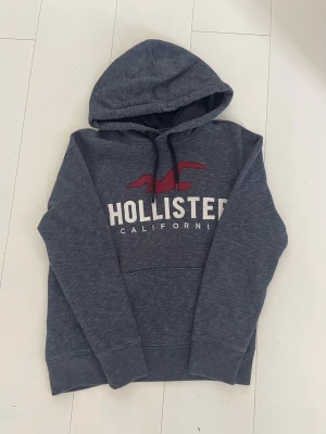 Mörkgrå Hollister hoodie med tryck - Mysig mörkgrå hoodie från Hollister. Perfekt för chill dagar. Mjuk bomullsblandning som känns skön mot huden. Ena snöret e lite sönder men funkar fortfarande lika bra. Jag är 160cm lång. Om man vill ha det lite stort så passar det för 150-155 om man vill ha det bra 156-163 om man vill ha den tajt 164-167.