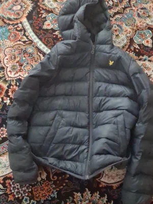 Svart pufferjacka från Lyle & Scott - Snygg svart pufferjacka från Lyle & Scott med huva och dragkedja framtill. Jackan har det klassiska Lyle & Scott-loggan i gult på bröstet och är vadderad för extra värme. Perfekt för kalla vinterdagar och enkel att matcha med olika outfits.