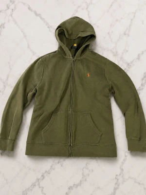 Olivgrön hoodie med dragkedja - Snygg olivgrön hoodie med dragkedja och huva. Klassisk design med kängurufickor framtill och en liten broderad logga på bröstet. Tillverkad i mjukt bomullsmaterial som känns skönt mot huden. Perfekt för en avslappnad och stilren look. De står xl men de är amerikanska storlekar så för bästa passform är den vid 170 cm men passar andra också. Vid frågor hör av dig och priset kan alltid diskuteras 