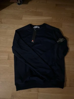 Mörkblå stickad tröja Stone Island - Snygg mörkblå stickad tröja från Stone Island med klassisk rund halsringning och ikonisk logotyp på ärmen. Tröjan har långa ärmar och ribbade muddar vid ärmslut och nederkant. Perfekt för en stilren och avslappnad look.