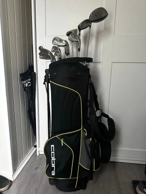 Golfset med Cobra bag och klubbor och skor - Säljer ett komplett golfset med svart och gul Cobra golfbag samt flera järnklubbor och drivers. Klubborna är i metall och inkluderar bland annat en Spalding wedge. Perfekt för dig som vill börja spela golf eller uppgradera din utrustning. Säljer också ett par golfskor