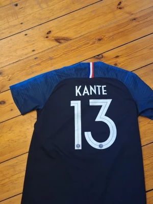 Blå fotbollströja Kante 13 - Snygg blå fotbollströja med KANTE och nummer 13 på ryggen. Tröjan har korta ärmar, mörkblå bas och blå detaljer på axlarna samt en liten fransk flagga i nacken. Perfekt för dig som vill stötta ditt lag på match eller träning.