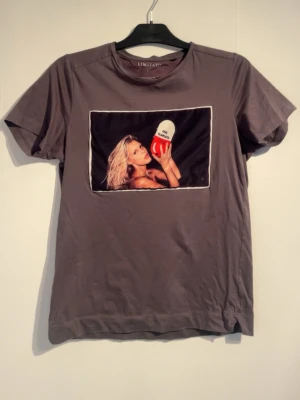 Brun t-shirt Limitato x Camilo Rios White - Snygg brun t-shirt från Limitato i samarbete med Camilo Rios White. Framsidan har ett stort tryck med en kvinna som håller i en rödvit kapsel med texten 'ART PILLS'. T-shirten har rund hals och är tillverkad i mjukt material.