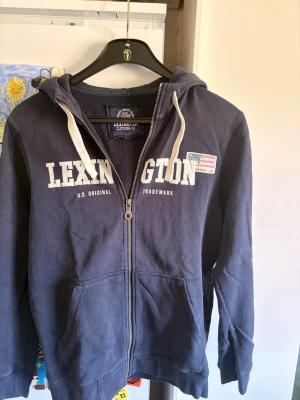 Marinblå zip-hoodie från Lexington - Marinblå hoodie med dragkedja från Lexington. Stor vit logga framtill och amerikansk flagga på bröstet. Justerbar huva med vita snören och två fickor fram. Tillverkad i mjukt bomullsmaterial, perfekt för en avslappnad stil.