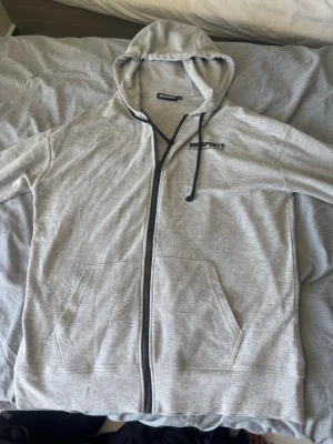 Grå zip hoodie från MM Sports - Snygg grå hoodie från MM Sports med dragkedja och svart tryck på bröstet. Hoodien har huva med snören och två stora fickor framtill. Perfekt för chill eller gymmet. Tillverkad i mjukt material för extra komfort.
