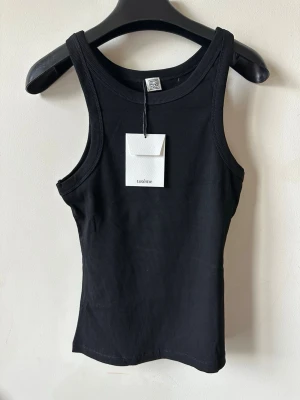 Svart ribbad linnetopp från Totême - Snygg svart ribbad linnetopp från Totême med klassisk rund halsringning och ärmlös design. Perfekt att styla till jeans eller kjol för en clean och enkel look. Materialet är mjukt och stretchigt för skön passform.