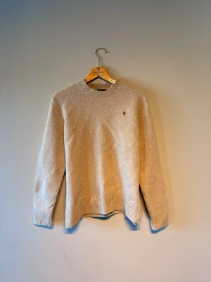 Beige stickad Morris tröja med brodyr - Snygg beige stickad tröja med rund hals och ribbade muddar. Tröjan har en liten broderad detalj på bröstet i form av en lilja. Perfekt för lager på lager och enkel att matcha med olika outfits.