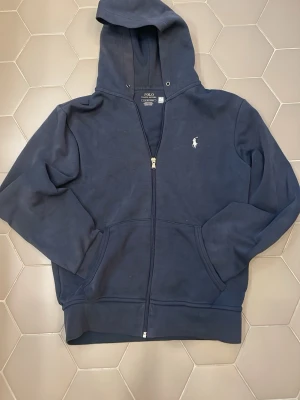 Marinblå hoodie från Polo Ralph Lauren - Snygg marinblå hoodie med dragkedja från Polo Ralph Lauren. Klassisk design med huva, fickor framtill och den ikoniska vita logon broderad på bröstet. Tillverkad i mjuk bomullsblandning som känns skön mot huden. Perfekt för en avslappnad och stilren look.