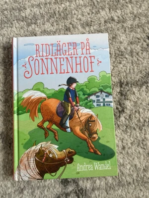 Ridläger på Sonnenhof - En färgglad barnbok med hårt omslag, skriven av Andrea Wandel. Omslaget visar en tecknad ryttare på en brun häst i en grön landskapsmiljö, med en gård i bakgrunden. Perfekt för dig som gillar hästar och äventyr.