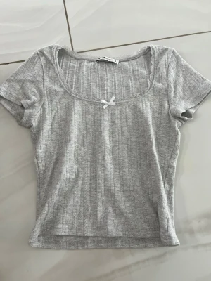 Grå ribbad topp med rosett - Supersöt grå ribbad topp från Sister med korta ärmar, rundad halsringning och liten vit rosett framtill. Toppen har diskret spetskant vid halsen och är perfekt för en chill och trendig look.