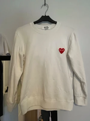 CDG pullover - Vit stickad tröja med rund hals och lång ärm. På bröstet finns ett litet rött hjärta med ögon, vilket ger en unik och lekfull detalj. Tröjan har ribbade muddar vid ärmslut och nederkant. Perfekt för dig som gillar enkel men cool stil.
