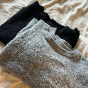 Grå och svart stickad tröja - Två stickade tröjor i mjukt material, en i ljusgrått och en i svart. Båda har rund halsringning och klassisk rak passform. Perfekta att matcha med jeans eller kjol för en enkel och stilren look.