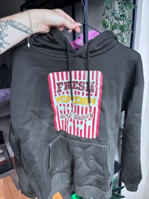Svart hoodie med popcorn-tryck - Cool svart hoodie med stor färgglad popcorn-tryck på bröstet där det står 'Fresh Popcorn Hot & Buttery'. Hoodien har huva med dragsko och en klassisk magficka framtill. Perfekt för dig som gillar unika och lekfulla plagg. Använd fåtal gånger. 